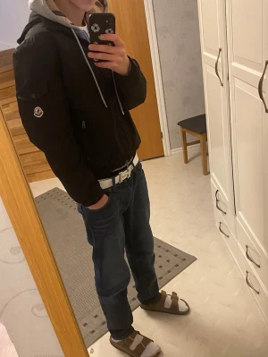 Svart Moncler dunjacka med huva - Svart Moncler dunjacka med huva och dragkedja framtill. Jackan har en snygg patch på ärmen, elastiska muddar och praktiska fickor. Materialet är polyester och dun, perfekt för kyliga dagar. Stilsäker och enkel att matcha med din outfit.