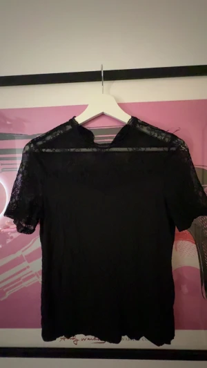 Svart blus med spetsdetaljer - Säljer en svart blus med korta ärmar och snygga spetsdetaljer vid axlar och hals. Blusen har en feminin och stilren look med transparent spets upptill och en mjuk, slät kropp. Perfekt att matcha med jeans eller kjol. Inga skavanker och fåsamma användningar 