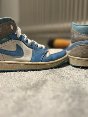 Nike Air Jordan 1 UNC Highs - Säljer ett par Nike Air Jordan 1 Highs i UNC-färgway med ljusblå och vita paneler, grå mockadetaljer och vit snörning. Skorna har en rund tå, platt sula och klassisk Jordan-logga på plösen. Perfekt för dig som gillar retro och streetwear.