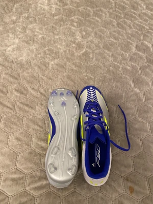 Adidas F50 blå och silver fotbollsskor - Säljer ett par Adidas F50 fotbollsskor i blått, silver och neon-gult. Skorna har snörning, platt sula med dobbar och en sportig design med de klassiska tre ränderna på sidan. Ovandelen är i syntetmaterial och insidan är fodrad i blått. Perfekt för dig som vill sticka ut på planen.