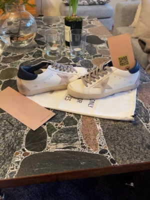 Golden Goose Super-Star sneakers, vit/blå - Golden Goose Super-Star sneakers i vitt skinn med blå häl och grå mockadetaljer. Ikonisk stjärna på sidan, grå snörning och platt sula. Snygg kontrast mellan färgerna och klassisk streetstil. Perfekt för dig som gillar italiensk design och coola detaljer.