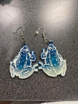 Blå glittriga grod örhängen  - Unika örhängen i form av grodor, med en snygg blå och vit gradient. Ena sidan har ett vitt konturmönster som framhäver grodans detaljer, medan baksidan är mer transparent och skimrande. Perfekt för dig som vill sticka ut med coola accessoarer.