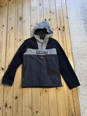 Napapijri anorak vindjacka - Snygg vindjacka från Napapijri i svart och grått med huva och halv dragkedja. Jackan har en stor logga på bröstet och norsk flagga, samt praktisk ficka framtill. Perfekt för dig som gillar streetwear och vill ha något som sticker ut. Hör av er vid frågor eller funderingar.