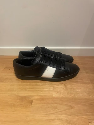 Svarta sneakers från Saint Laurent - Säljer ett par svarta sneakers från Saint Laurent med en vit sidopanel och svarta skosnören. Skorna har rund tå, platt sula och är tillverkade i skinn. Snygg och stilren design med diskret logga på sidan och hälen.