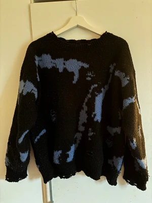 Svart stickad tröja med blått mönster - Cool svart stickad tröja med blått abstrakt mönster. Tröjan har en loose passform, ribbad hals och muddar samt långärmade ärmar. Detaljer med sliten look och hål ger en edgy vibe. Perfekt för dig som gillar streetstyle och unika plagg.