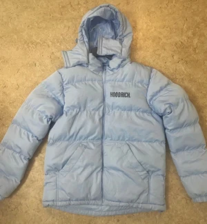 Ljusblå pufferjacka från Hoodrich - Säljer en ljusblå pufferjacka från Hoodrich med stor logga på ryggen och mindre logga på bröstet. Jackan har huva, dragkedja och två stora fickor framtill. Perfekt för kalla dagar när du vill ha en chill och snygg look.