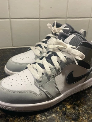 Nike Jordan 1 Mid i grå och vit - Nike Jordan 1 Mid sneakers i en clean färgway med grå och vita paneler, svart swoosh och vita snören. Skorna har en rund tå, platt sula och klassisk snörning. Ovandelen är i skinn och textil, med vadderad krage för extra komfort. Perfekt för dig som gillar streetwear och snygga sneakers.