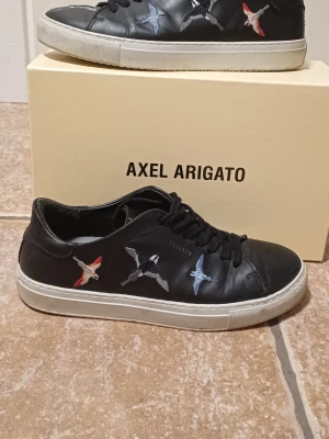 Svarta sneakers från Axel Arigato - Svarta sneakers från Axel Arigato med broderade fåglar i blått, grått och rött på sidorna. Tillverkade i skinn. 