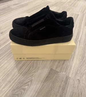 Svarta sneakers från Axel Arigato - Snygga svarta sneakers från Axel Arigato med minimalistisk design och diskret logga i guld på sidan. Skorna har en rund tå, platt sula och är tillverkade i mocka för en lyxig känsla. Perfekta för dig som gillar stilrena och moderna skor.