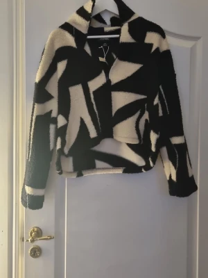 Svartvit teddyjacka från Monki XXS - Säljer en unik fluffig teddyjacka från Monki i storlek XXS. Jackan har ett  grafiskt mönster i svart och vitt, med en quarter dragkedja framtill och hög krage. Materialet är mjukt och cozy polyester, perfekt för att sticka ut i höst och vinter.