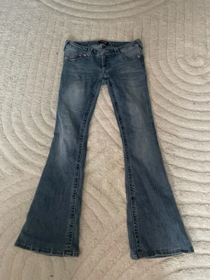True Religion bootcut jeans blå, stl 27 - Säljer ett par klassiska blå bootcut jeans från True Religion med ikoniska bakfickor och snygga kontrastsömmar. Jeansen har låg midja och är tillverkade i jeansmaterial med en lätt tvättad look. Perfekta för dig som gillar en avslappnad men trendig stil.  Använda två gånger💕