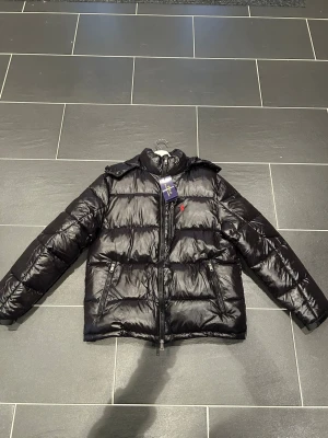 Svart pufferjacka från Polo Ralph Lauren - Svart pufferjacka från Polo Ralph Lauren med glansig finish, röd logga på bröstet och två dragkedjeförsedda fickor. Jackan har hög krage och quiltad design som ger en cool och sportig vibe. Perfekt för kalla dagar.