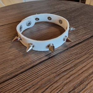 Vit punk choker med silverfärgade nitar - Armbaand punk. Cool vit choker med flera silverfärgade konformade nitar och tryckknappar. 24 cm långt. Bredd 1,5 cm. Armbandet är tillverkat i syntetmaterial Pu läder och har en edgy vibe som passar perfekt till streetwear eller alternativa stilar.