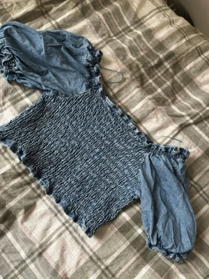 Blå smockad blus från Levi's - Säljer en blå smockad blus från Levi's i storlek L. Toppen har puffiga korta ärmar med resår och en smockad kropp som ger en snygg och figurnära passform. Materialet är mjukt och tillverkat av TENCEL™ lyocell, vilket gör den både skön och hållbar. Perfekt för dig som gillar denimlook!