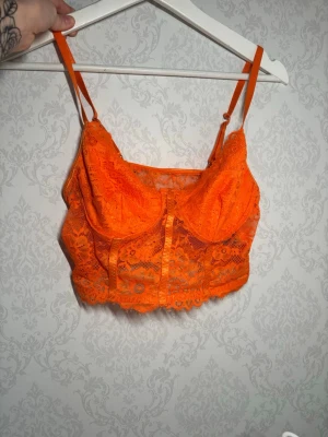 Orange spets korsett-topp - Säljer en orange korsett-topp med tunna axelband och vackert blommigt spetsmönster. Toppen har dekorativa band framtill och en scallop-kant längst ner. Perfekt för dig som gillar färgstarka och detaljerade plagg.