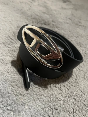 Svart vintage diesel bälte  - Snyggt svart bälte med ett ovalt, silverfärgat spänne som har en unik design. Bältet är stilrent och passar perfekt till jeans eller kostymbyxor. Materialet känns som skinn och spännet ger en cool touch till outfiten.