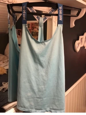 Ljusblå linnetopp med Just Do It-band - Säljer en ljusblå linnetopp med breda blå axelband där det står 'Just Do It.' i vitt. Toppen har en enkel och rak form, är ärmlös och tillverkad i mjuk bomull. Perfekt för dig som gillar sportiga detaljer och vill ha en chill look.