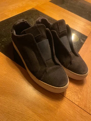 axel arigato laceless - Säljer ett par svarta sneakers från axel arigato med stilren design och bred elastisk rem över vristen. Skorna har en vit platt sula och är tillverkade i mjuk mocka som ger en schysst look. Perfekt för dig som gillar minimalistiska och bekväma skor.