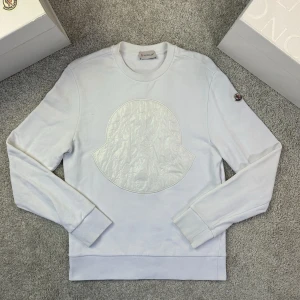 Moncler Sweatshirt - Säljer en stilren vit sweatshirt från Moncler med stort, ton-i-ton logomärke på bröstet. Tröjan har rund hals, långa ärmar och ribbade muddar. Perfekt för dig som gillar exklusiva märken och enkel design. Storlek S