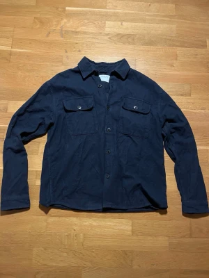 Mörkblå overshirt från Selected Homme - Snygg mörkblå overshirt från Selected Homme med klassisk krage och två bröstfickor med knappar. Jackan har långärmad design och är tillverkad i mjuk bomull, perfekt för lager-på-lager. Stängs med knappar framtill och har en clean look. Passar med allt och lätt att stylea med