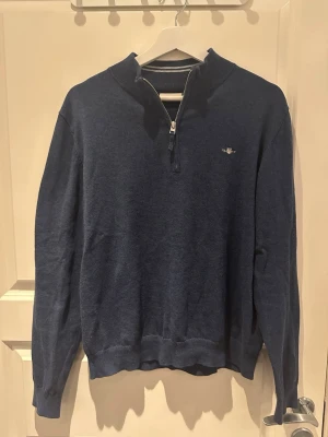 Blå half zip tröja från GANT - Snygg mörkblå tröja från GANT med half zip och liten broderad logga på bröstet. Tröjan har ribbad krage med vita ränder, långa ärmar och är gjord i mjuk bomull. Perfekt för dig som gillar klassisk stil med en sportig touch.