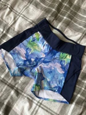Blommiga träningsshorts från SOC - Säljer ett par snygga träningsshorts från SOC i storlek M. De har ett blommigt mönster i blått, grönt och vitt med marinblå midja och sidor. Materialet är syntet och designen är tight med hög midja, perfekt för gym eller löpning.