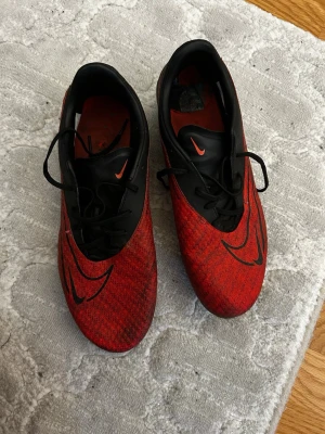 Röda och svarta Nike fotbollsskor - Säljer ett par snygga fotbollsskor från Nike i rött och svart med coolt mönster och svarta detaljer. Skorna har snörning och rund tå, samt en platt sula med dobbar för bra grepp på planen. Perfekt för dig som vill sticka ut på matchen!
