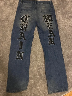 Blå jeans med tryck 'CHAIN WEAR' - Unika blå jeans med stort tryck i svart och vitt på baksidan där det står 'CHAIN' på ena benet och 'WEAR' på det andra. Jeansen har raka ben och klassisk femficksdesign. Materialet är robust denim och de har en cool streetstil.