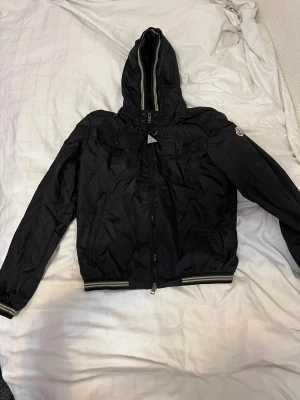 Moncler Vindjacka - Svart vindjacka från Moncler med huva och dragkedja framtill. Jackan har två bröstfickor med knappar, ribbade muddar med ränder och justerbar dragsko i huvan. Tillverkad i lätt polyester, perfekt för blåsiga dagar. Storlek 1 som motsvarar S. Passar även XS. Pris kan diskuteras vid snabb affär.