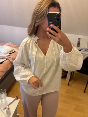 Vit blus 100% bomull - Luftig och bekväm blus i vit, lätt strukturerad bomull. Modellen har lång ärm med resår i ärmsluten, v-ringad hals med knytband samt en avslappnad passform som faller fint. Perfekt både till vardag och mer uppklädda tillfällen. Mycket skön att bära och lätt att matcha.Fin blus helt felfri. Den är 100% bomull!! 