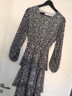 Leopardmönstrad långklänning med volanger - Snygg långklänning med svart och grått leopardmönster. Klänningen har långa ärmar, markerad midja med snörning och flera lager volanger i kjolen för extra rörelse. Tillverkad i ett lätt och luftigt syntetmaterial som ger en cool vibe.