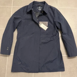 Mörkblå trenchcoat från UBR L - Snygg mörkblå trenchcoat från UBR med klassisk krage och knappstängning. Jackan har stilren design, raka linjer och är tillverkad i ett tekniskt syntetmaterial som står emot väder. Perfekt för dig som gillar minimalistisk och modern stil.