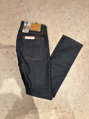 Helt nya Nudie Jeans mörkblå jeans - Mörkblå jeans från Nudie Jeans Co, tillverkade i 100% ekologisk bomull. Klassisk femficksdesign med orange kontrastsömmar och Nudie-logga på bakfickan. Slimmad passform och snyggt skinnpatch bak i midjan. Helt nya med alla ”taggar”