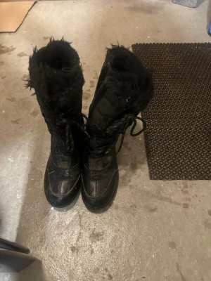 Svarta vinterstövlar med fuskpäls - Säljer ett par svarta vinterstövlar från Lakeforest i storlek 39. Skorna har snörning framtill, fuskpäls runt skaftet och är tillverkade i syntetmaterial. Perfekta för kalla dagar med sin höga modell och fluffiga insida.