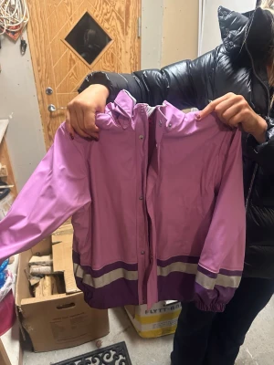 Lila regnjacka med reflexdetaljer - Säljer en lila och ljuslila regnjacka från Didriksons med huva och reflexdetaljer på ärmar, nederkant och huva. Jackan har tryckknappar framtill, elastiska muddar och trycket '1913' på ryggen. Perfekt för regniga dagar och snygg att matcha med boots.