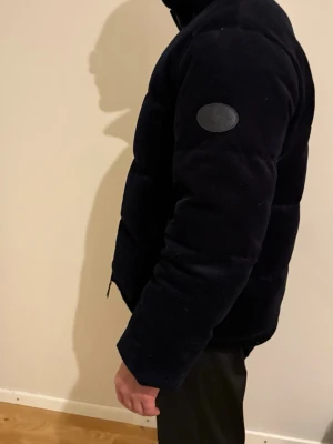 Svart manchester pufferjacka L - Säljer en svart pufferjacka i manchester med hög krage och dragkedja framtill. Jackan har en stilren look med diskret patch på ärmen och breda paneler. Perfekt för dig som vill ha en varm och trendig jacka i vinter.