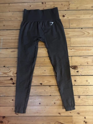 Mörkgrå seamless leggings från Gymshark - Säljer ett par bruna seamless leggings från Gymshark med hög midja och diskret strukturmönster längs benen. Stretchigt material som sitter tight och följer kroppen. Perfekta för träning eller chill. Gymshark-logga på höften och vid benslut.