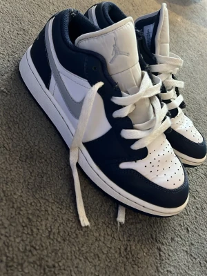Nike Air Jordan 1 Low marinblå/vit - Säljer ett par Nike Air Jordan 1 Low i marinblått och vitt med grå swoosh. Skorna har klassisk snörning, perforerad tåbox och Air Jordan-logga broderad på hälen. Tillverkade i skinn och syntetmaterial för en clean och sportig look. Knappt använda.
