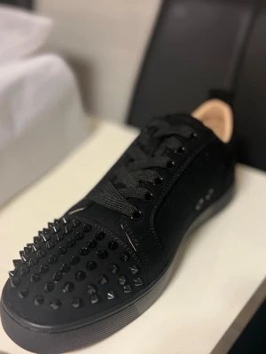 Christian Louboutin svarta sneakers med nitar - Säljer ett par svarta sneakers från Christian Louboutin med mattsvarta nitar på tån och den ikoniska röda sulan. Skorna har snörning, rund tå och är tillverkade i mocka och skinn. Perfekta för dig som vill sticka ut med en edgy och lyxig vibe.