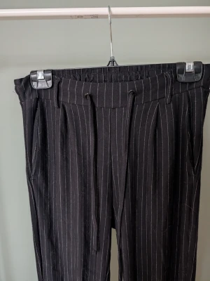 Svarta randiga byxor med snörning - Säljer ett par svarta byxor med vita tunna ränder och snörning i midjan. Byxorna har hög midja, resår i midjan och smal passform. Materialet känns mjukt och stretchigt, perfekt för en chill och snygg look.