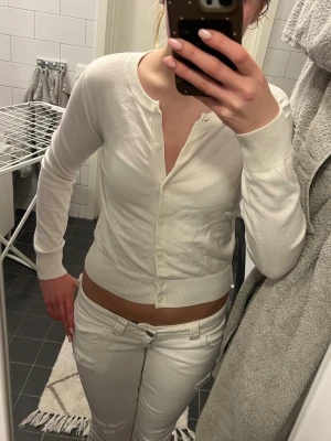 Vit kofta med knappar - Säljer en enkel och stilren vit kofta med rund hals och knappar framtill. Tröjan har lång ärm och ribbade muddar vid ärmslut och nederkant. Perfekt att matcha med jeans eller kjol för en clean look.