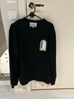 Svart sweatshirt från Casablanca M - Svart sweatshirt från Casablanca med färgglad grafisk print på bröstet och ryggen. Tröjan har rund hals, långa ärmar och en relaxed passform. Perfekt för dig som gillar streetwear och vill sticka ut lite extra.