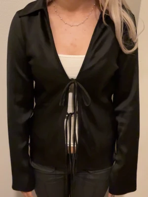 Svart omlottblus i satin med snörning - Säljer en svart omlottblus i glansigt satinliknande material. Blusen har långa ärmar med slits vid ärmslut och knyts framtill med två snören. Snygg v-ringning och figurnära passform. Perfekt att styla med ett linne under💗