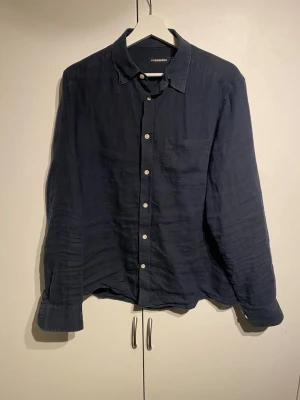 Mörkblå linneskjorta från J.Lindeberg - Snygg mörkblå skjorta från J.Lindeberg i 100% linne. Klassisk krage, vita knappar framtill och en bröstficka. Storlek M, avslappnad passform. Välanvänd men i fint skick. Nypris 1400kr. 