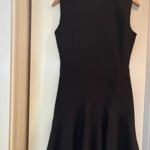 Svart ärmlös klänning med volang - Svart, ärmlös klänning med rund hals och volang nedtill. Klänningen har en figurnära passform upptill och en lätt utsvängd kjol. Materialet är mjukt och slätt, troligen bomull eller polyester. Dragkedja baktill för enkel påklädning. Perfekt för dig som gillar stilren och enkel design.