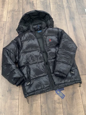 Svart pufferjacka från Polo Ralph Lauren - Säljer en svart pufferjacka från Polo Ralph Lauren med huva och dragkedja. Jackan har en klassisk röd logga på bröstet, två fickor med dragkedja och justerbar nederkant. Materialet är glansigt och fylligt, perfekt för kalla dagar. Kvitto finns! Köpte en ny jacka så jag behöver inte denna mer? Pris kan diskuteras vid snabb affär 
