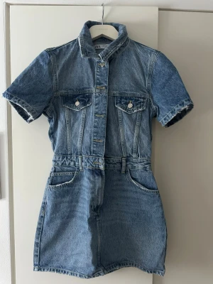 Jeans byxdress från Zara, blå denim - Säljer en snygg blå jeans byxdress från Zara med korta ärmar, krage och knappar framtill. Byxdressen har två bröstfickor med lock och knapp samt sidofickor. Tillverkad i klassisk denim med markerad midja och cool vintagekänsla.
