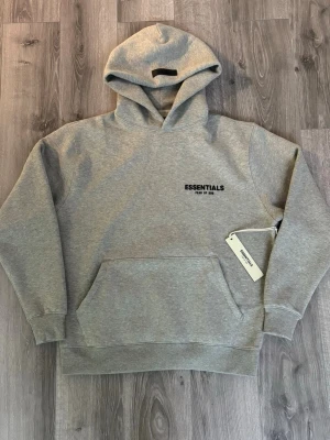 Essentials Fear of God Hoodie – Grå  - Beskrivning: Säljer en Essentials Fear of God hoodie i grå färg. Klassisk och stilren modell med Essentials-logga på bröstet. Tjock och bekväm kvalitet med avslappnad passform. Storleken är xs men men om du har st S Eller M så kommer Deta vara perfect fit för dig, annars lite baggy  Hoodien är i mycket bra skick och kommer med originaltag kvar (se bild). Perfekt både till vardags och streetwear-outfits.  ✔️ Populär Essentials-modell ✔️ Bekväm & hög kvalitet  Tar emot bud,köpt är köpt