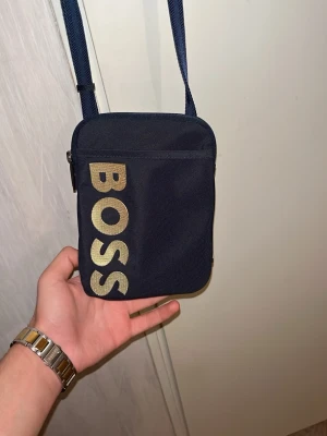 Svart axelväska från BOSS - Säljer en stilren svart axelväska från BOSS med justerbar rem och stort guldfärgat BOSS-tryck på framsidan. Väskan har dragkedja upptill och ett rymligt huvudfack. Perfekt för att bära det viktigaste och ger en clean look. Ligger på 800k-900kr som ny 