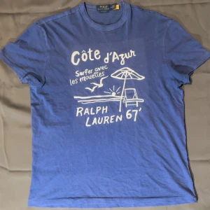 Ralph Lauren T Shirt - Blå t-shirt från Polo Ralph Lauren med vit tryckt grafik på framsidan som visar strandmotiv, parasoll och texten 'Côte d'Azur'. Tröjan är knappast använd och i nytt skick. Om du har fler frågar är de bara att kontakta mig.                                         